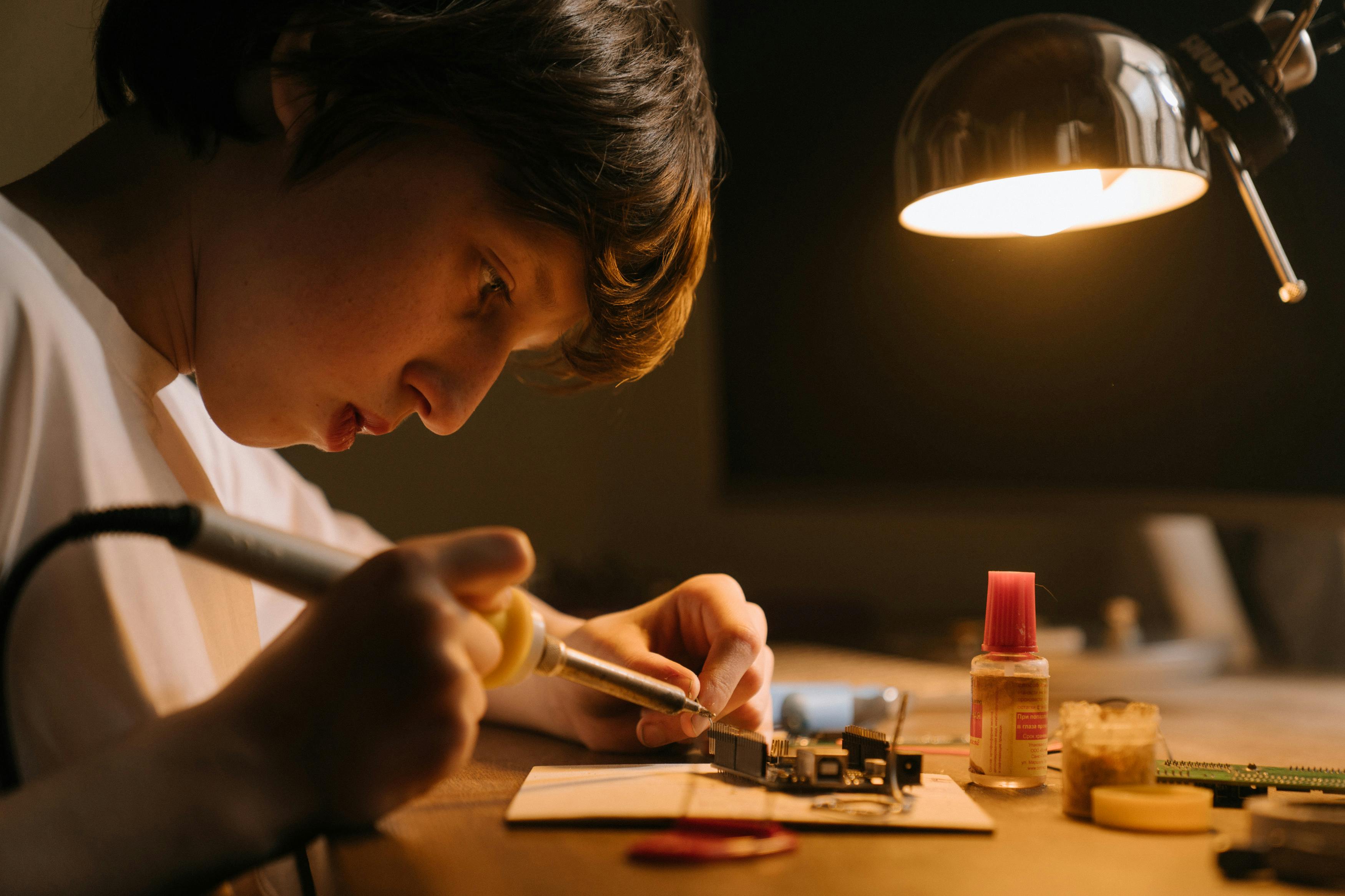 Engineer soldering a circuit board at a workbench under warm desk light.